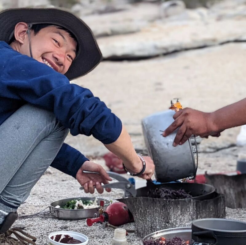 In the image, a person wearing a blue sweater and a hat is smiling while cooking outdoors. They are crouched near a portable stove, where someone is pouring food from a pot into a pan. Other cooking utensils and ingredients are scattered around the cooking area, suggesting a meal preparation scene in a camping or outdoor setting.